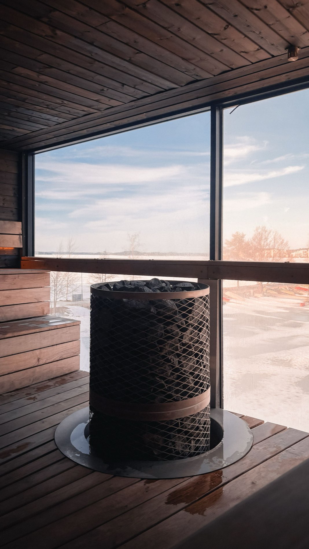 Inside a warm cedar sauna with a stone heater, looking out through a large window over a frozen snow-covered lake